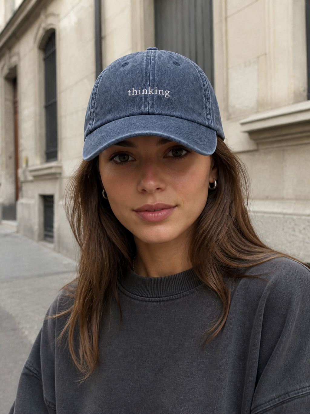 A woman in a charcoal sweatshirt wearing the washed navy 'thinking' cap on a city street in front of a stone building.