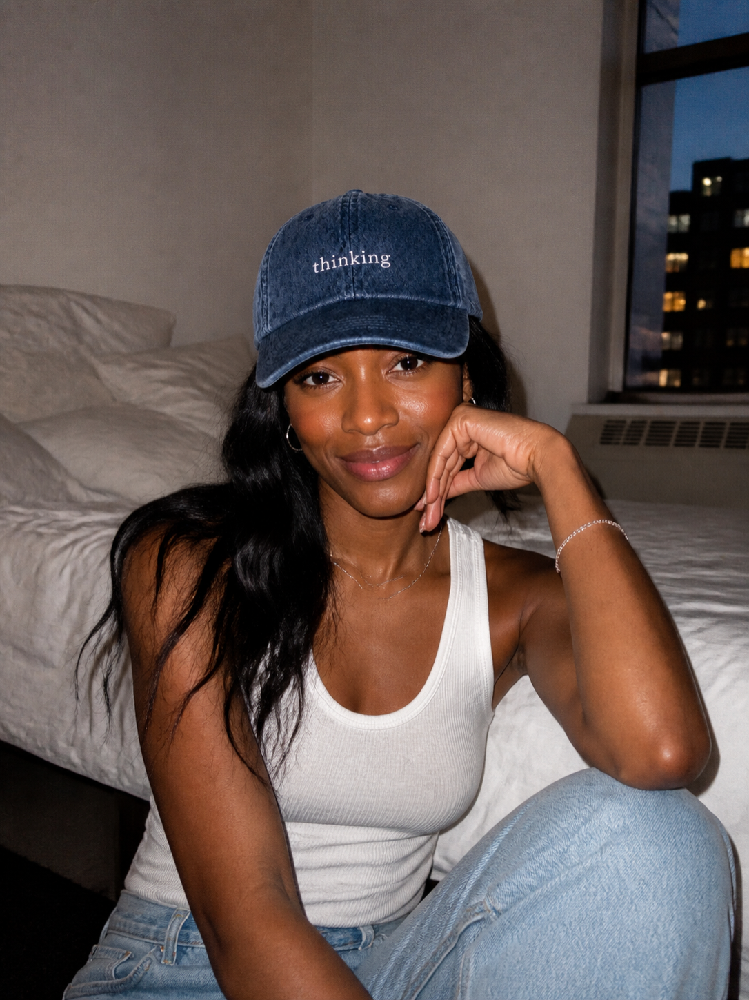 A woman in a white tank top wearing the washed navy 'thinking' cap, seated indoors by a window at dusk.