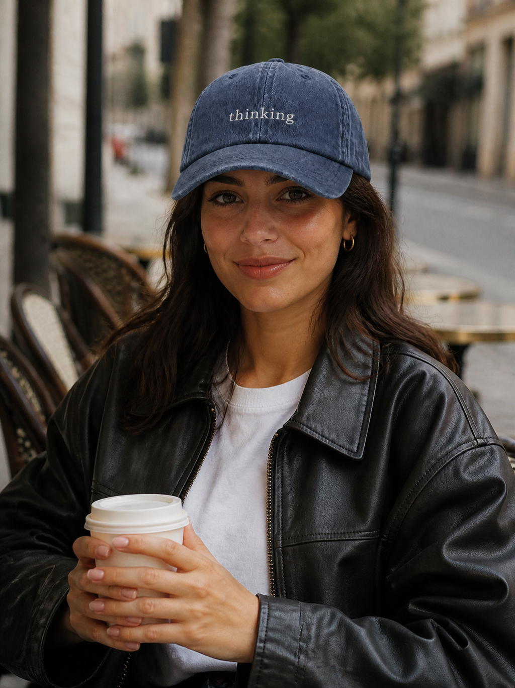 A woman in a black leather jacket holding a coffee cup and wearing the washed navy 'thinking' cap at a cafe terrace.