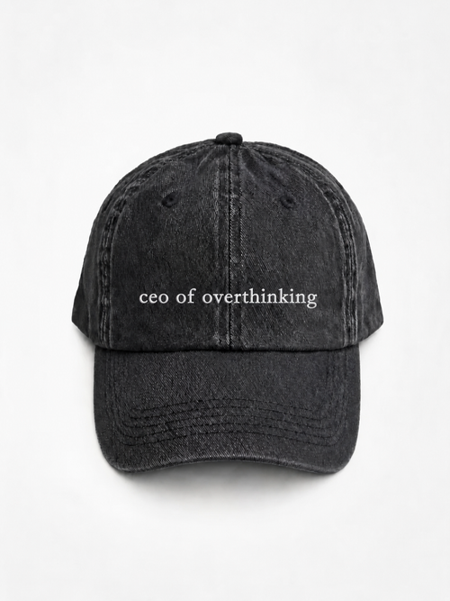 The washed black 'ceo of overthinking' cap photographed flat and front-on, showing the white embroidered lettering across the front panel.
