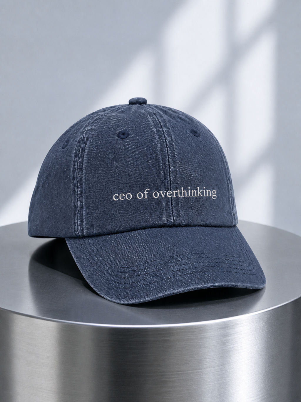 The washed navy 'ceo of overthinking' cap on a brushed steel pedestal in soft window light.