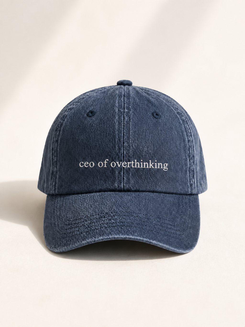 The washed navy 'ceo of overthinking' cap standing front-on against a warm cream backdrop.