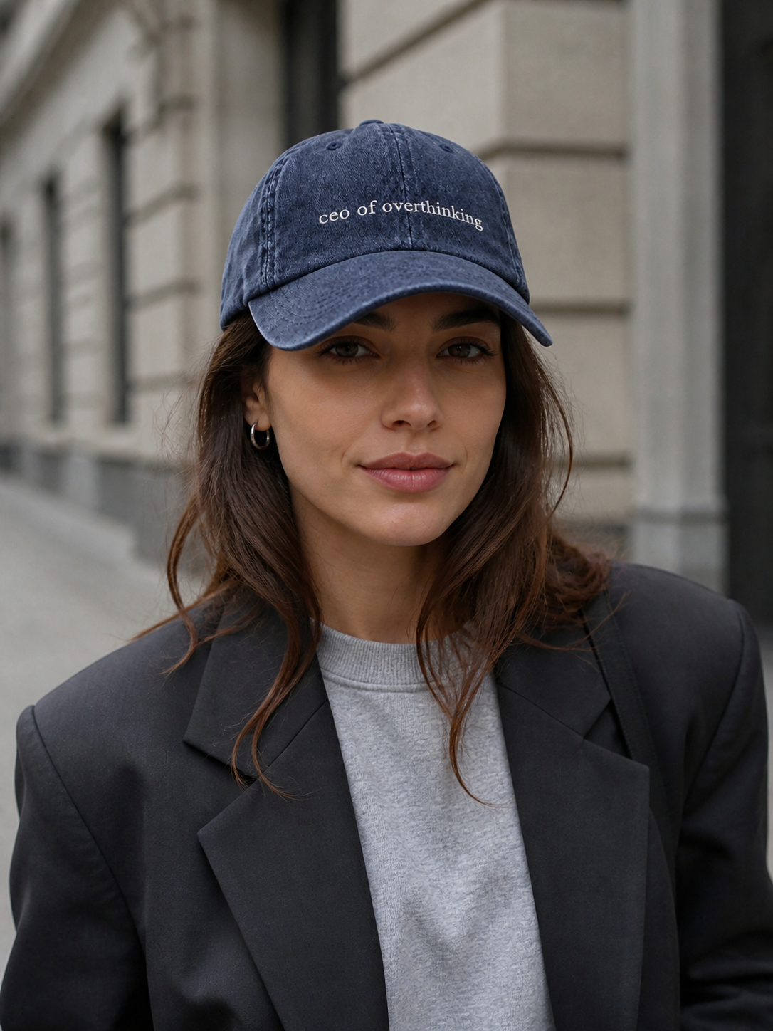 A woman in a dark blazer and grey sweatshirt wearing the washed navy 'ceo of overthinking' cap on a city street.