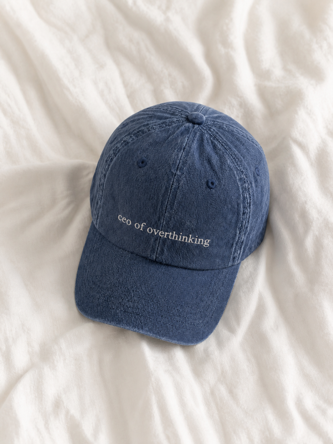 The washed navy 'ceo of overthinking' cap lying on rumpled cream bedsheets.