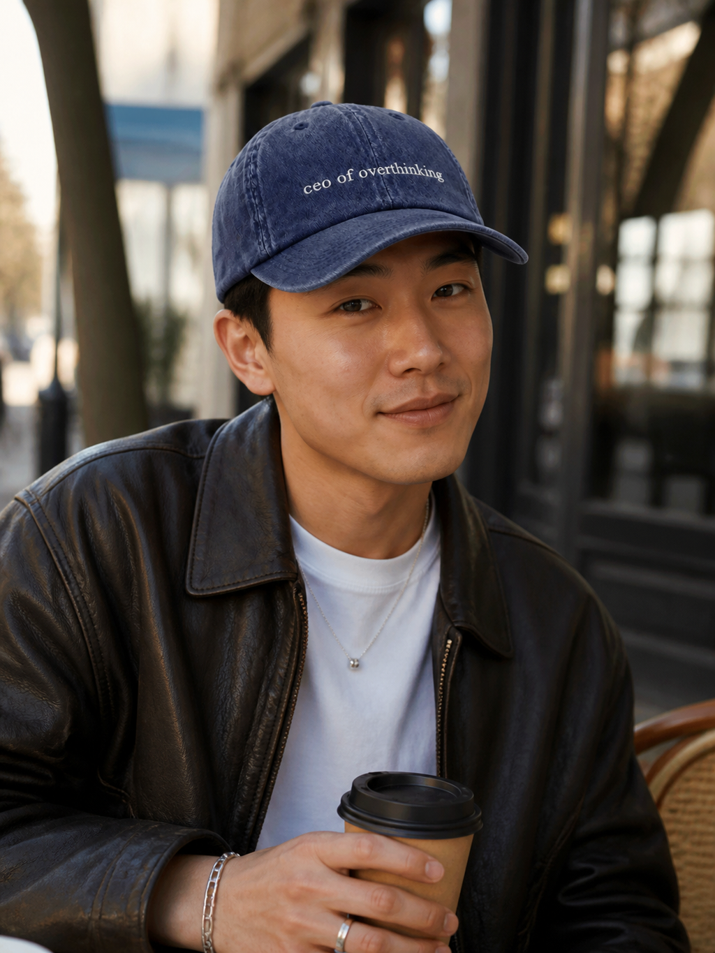 A man in a leather jacket wearing the washed navy 'ceo of overthinking' cap and holding a takeaway coffee on a cafe terrace.