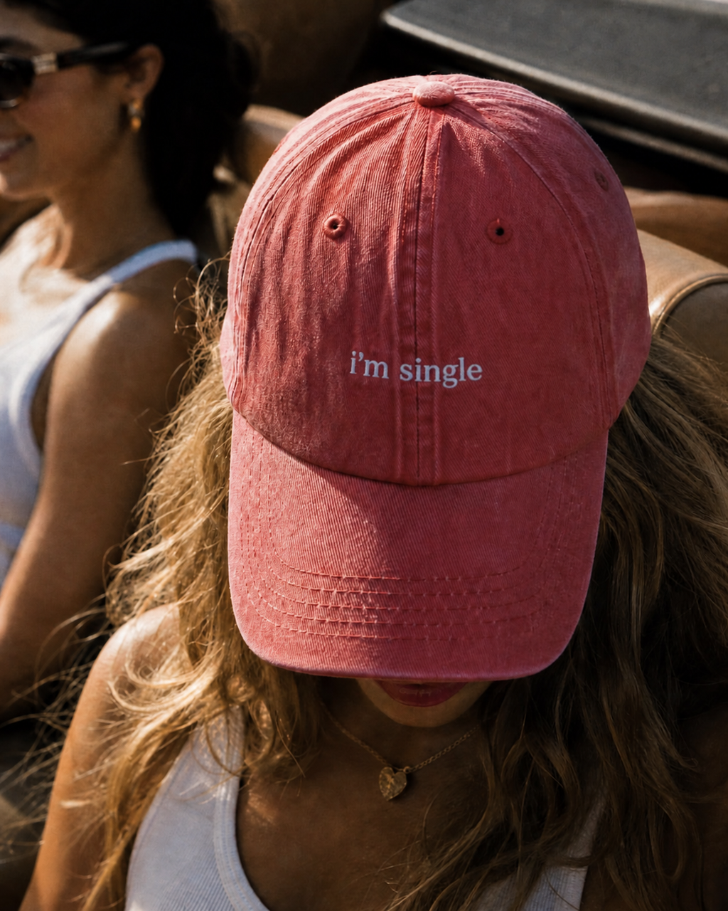 A dusty rose pink baseball cap with the text 'i'm single' embroidered in white on the front, worn by a woman in an outdoor setting.