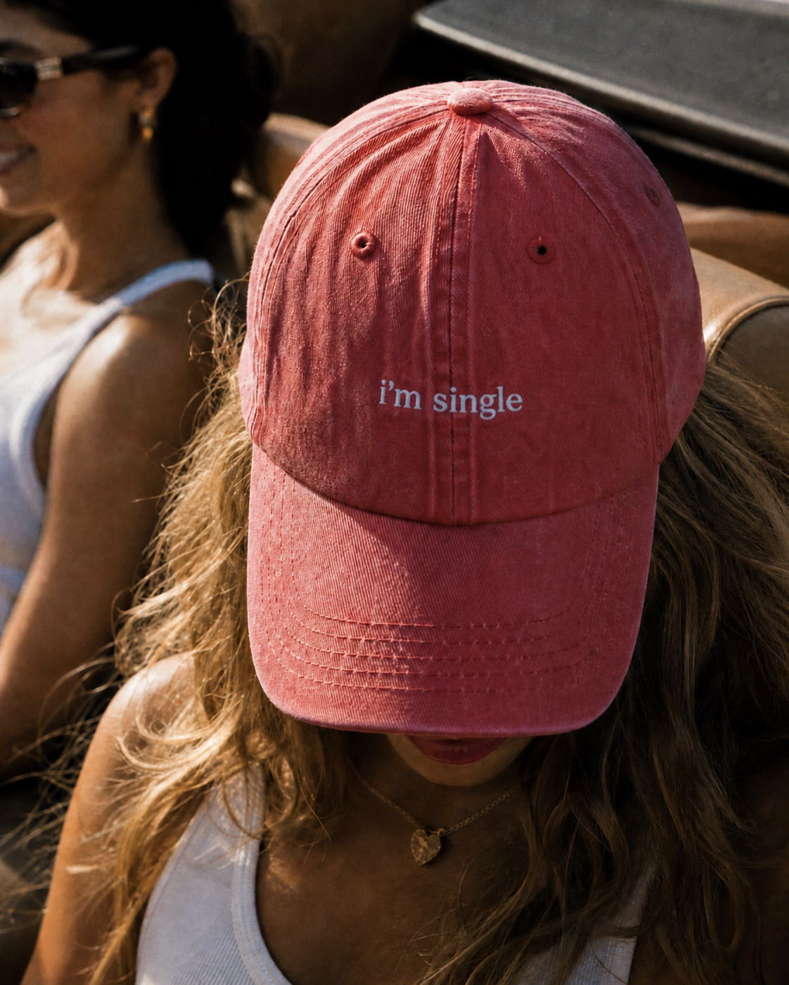 A dusty rose pink baseball cap with the text 'i'm single' embroidered in white on the front, worn by a woman in an outdoor setting.