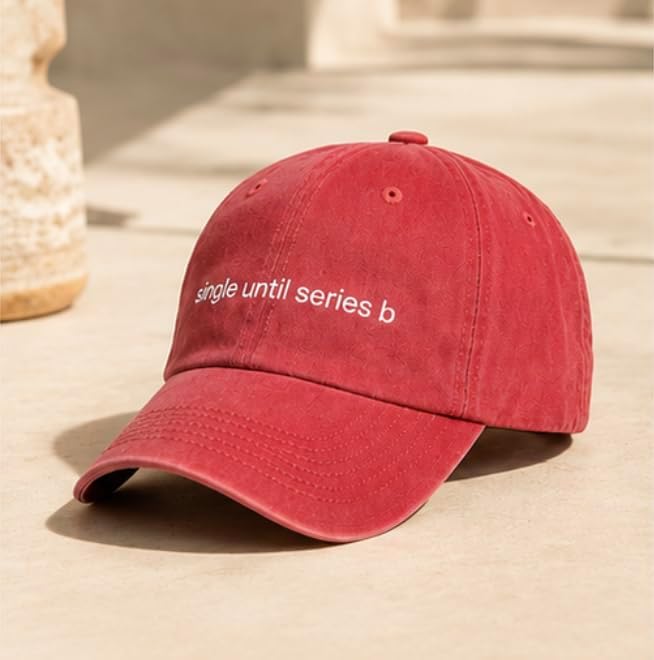 A red baseball cap with the phrase 'single until series b' embroidered in white on the front, resting on a beige stone surface.