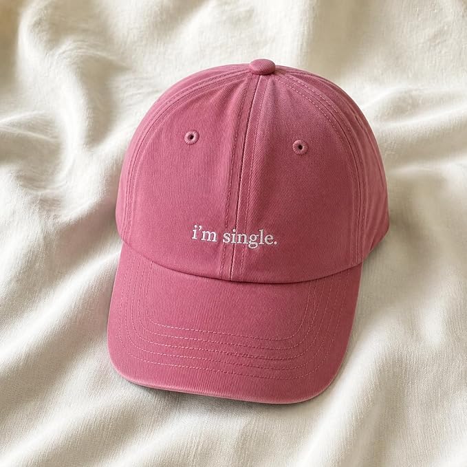 A dusty pink baseball cap with the phrase 'i'm single.' embroidered in white on the front, resting on a soft, draped cream-colored fabric background.