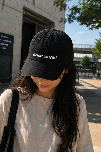 A woman outdoors wearing a black baseball cap with the word 'funemployed' embroidered in white across the front, paired with a light beige t-shirt and a black shoulder bag.