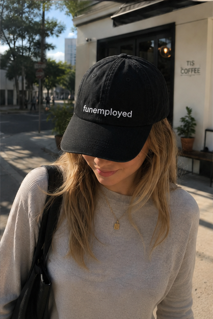 A woman wearing a black baseball cap with the text 'funemployed' embroidered in white on the front, photographed outdoors on a city street.