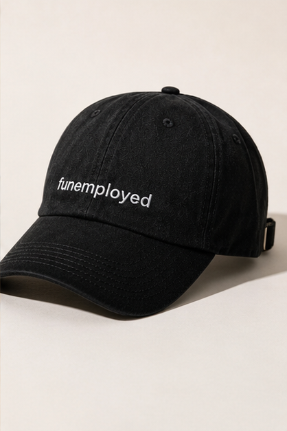 A black cotton baseball cap with the word 'funemployed' embroidered in white on the front, photographed on a clean, light beige background.