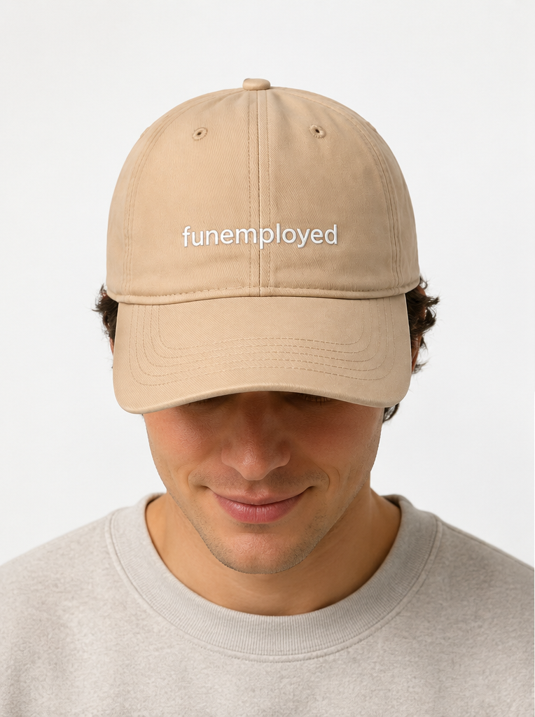 A man wearing a beige baseball cap with the word 'funemployed' embroidered in white on the front, paired with a light gray crewneck sweatshirt against a plain light gray background.