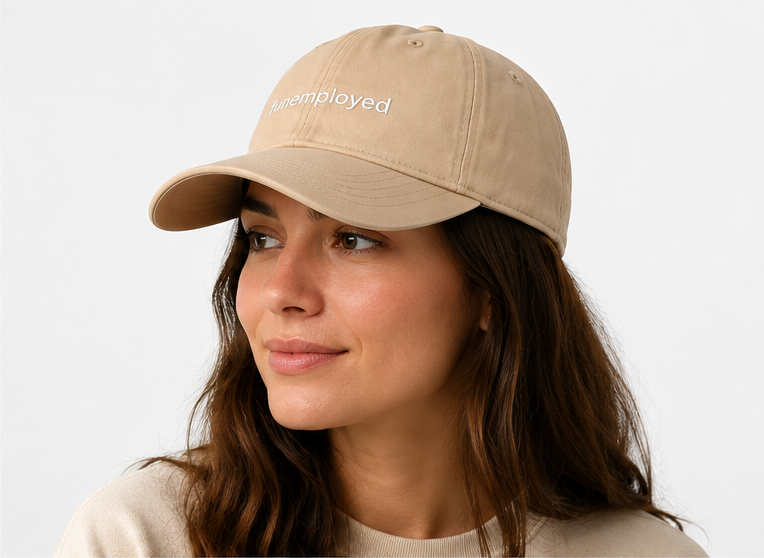 A woman wearing a beige baseball cap with the word 'unemployed' embroidered in white on the front, shown against a plain light gray background.