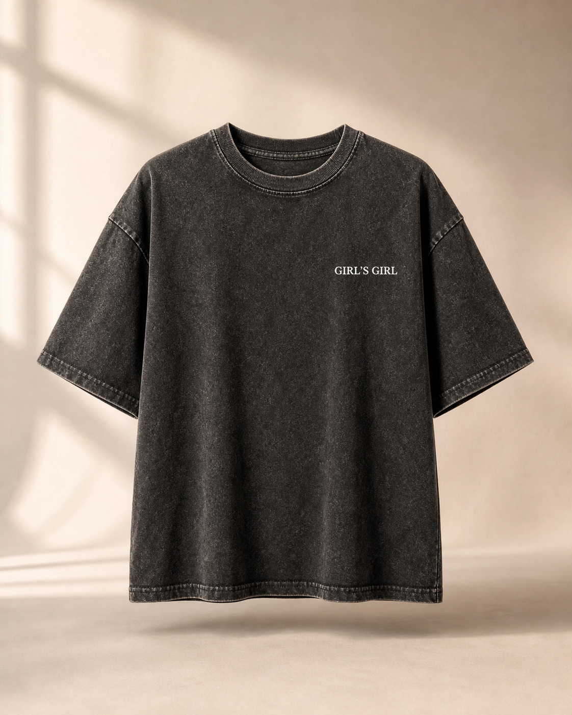 Washed black oversized t-shirt with GIRL'S GIRL printed in small white serif type on the left chest, against a warm beige studio backdrop with soft window light.