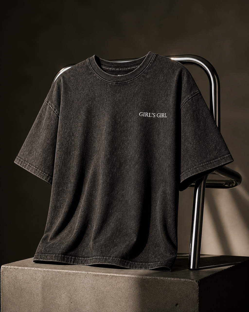 Washed black GIRL'S GIRL t-shirt draped over a chrome tubular chair on a concrete plinth, lit against a dark studio background.