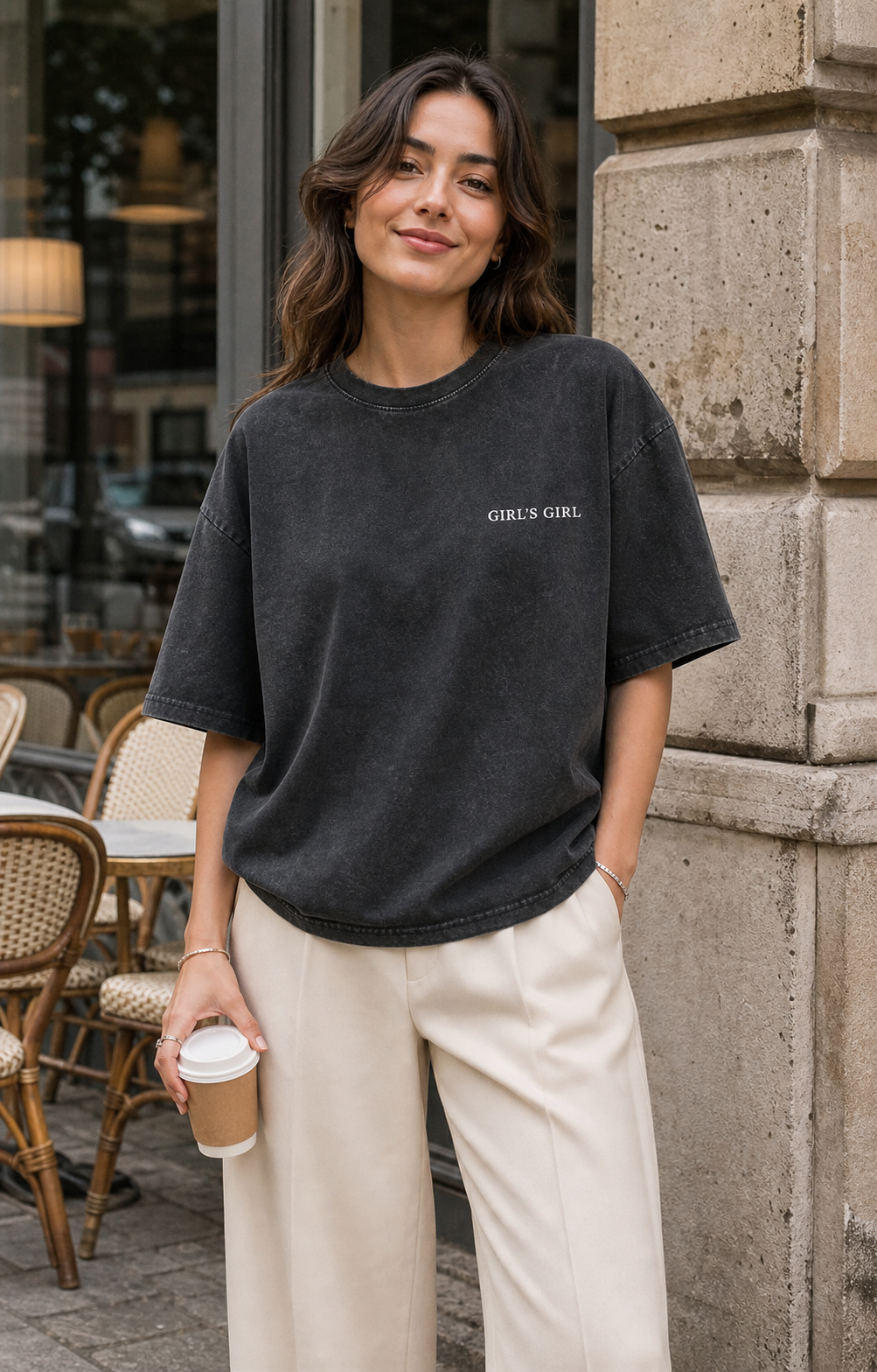 A woman outside a cafe wearing an oversized washed black GIRL'S GIRL t-shirt with cream wide-leg trousers, holding a takeaway coffee cup.