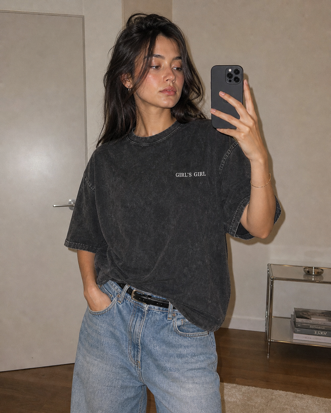 A woman taking a mirror selfie in an oversized washed black GIRL'S GIRL t-shirt tucked into light blue jeans with a black belt.