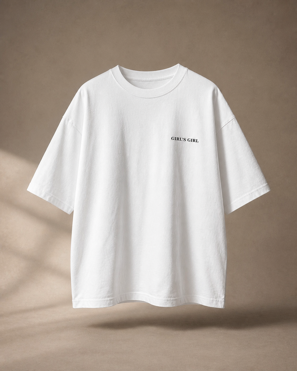 White oversized t-shirt with GIRL'S GIRL printed in small black serif type on the left chest, against a warm taupe studio backdrop with soft window light.