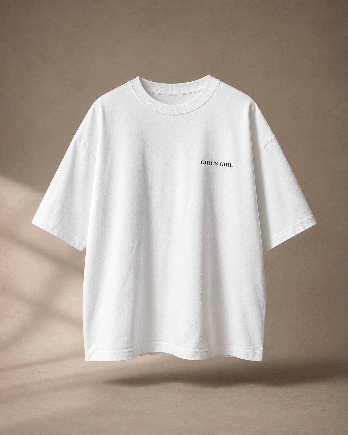 White oversized t-shirt with GIRL'S GIRL printed in small black serif type on the left chest, against a warm taupe studio backdrop with soft window light.