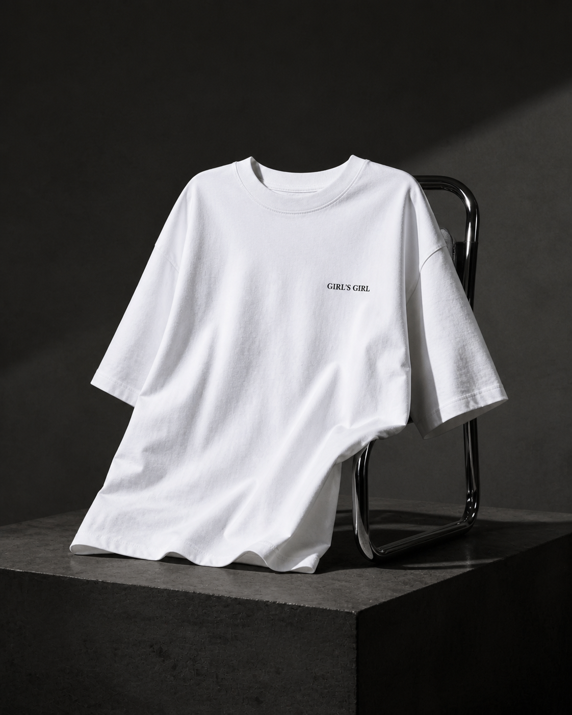 White GIRL'S GIRL t-shirt draped over a chrome tubular chair on a dark concrete plinth, lit against a charcoal studio background.