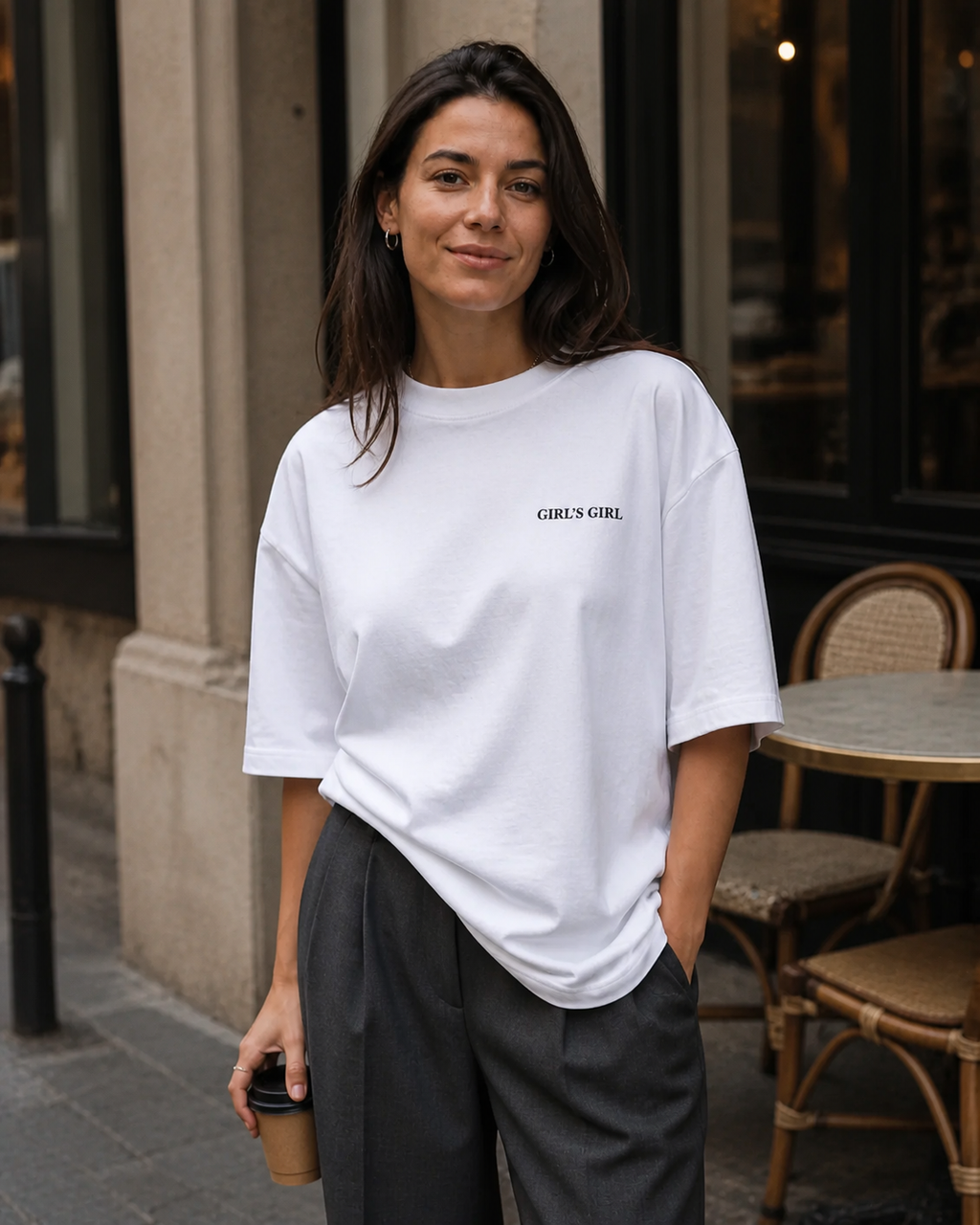 A woman outside a cafe wearing an oversized white GIRL'S GIRL t-shirt with charcoal pleated trousers, holding a takeaway coffee cup.