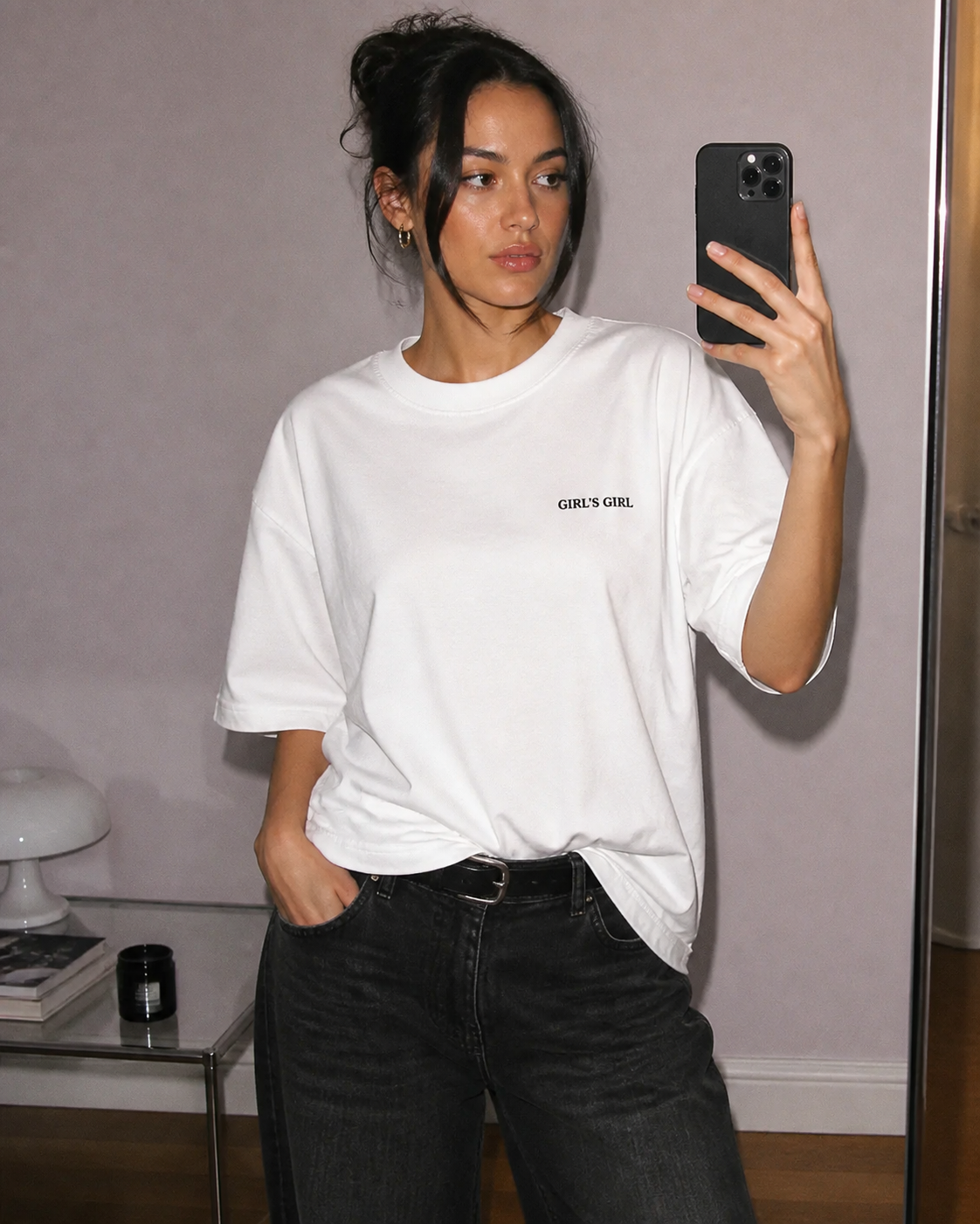 A woman taking a mirror selfie in an oversized white GIRL'S GIRL t-shirt tucked into black jeans with a leather belt.