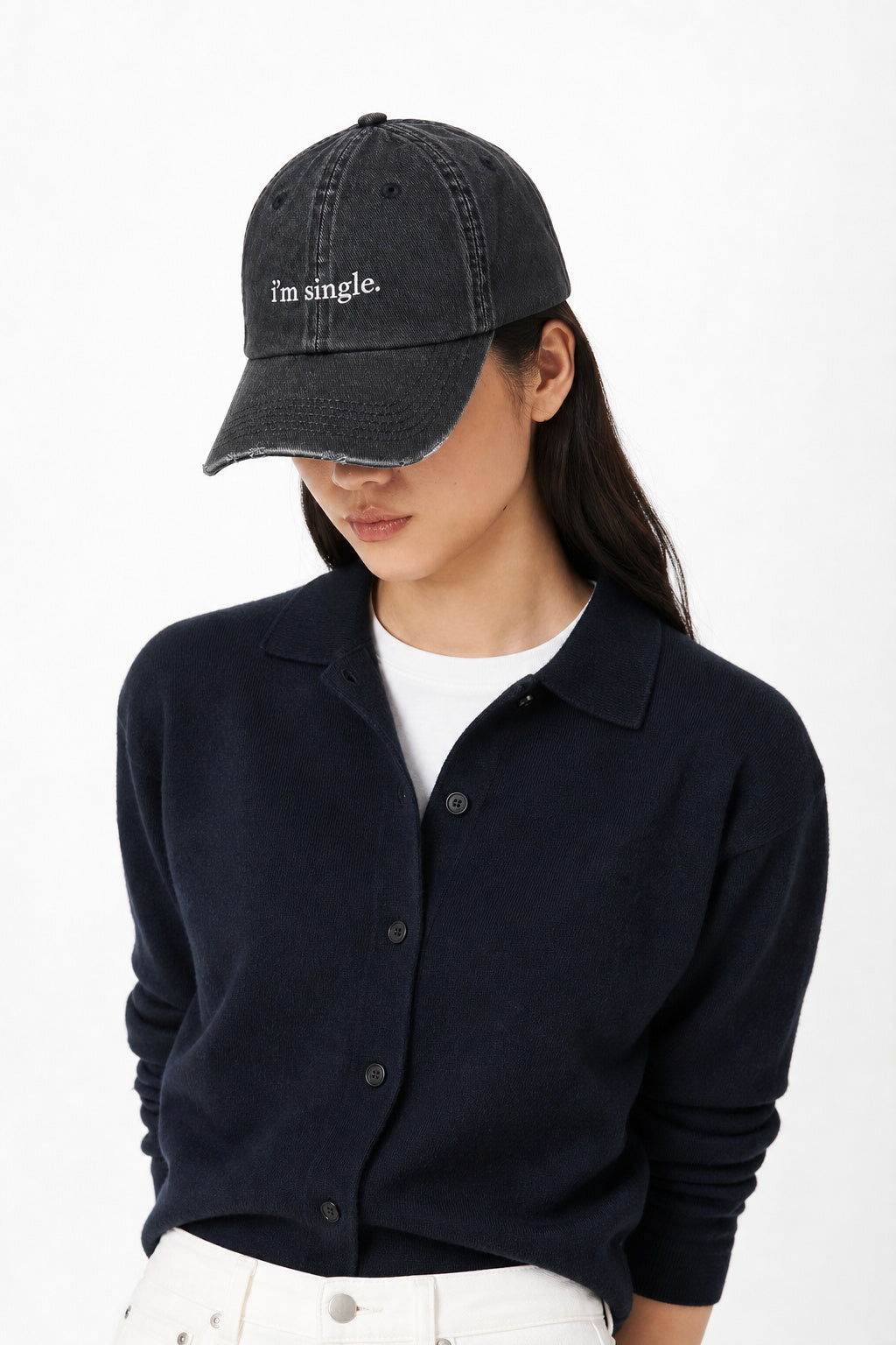 A woman wearing a washed black baseball cap with the embroidered text 'I'm single.' in white, styled with a dark navy button-up cardigan over a white t-shirt and white pants, against a plain white background.