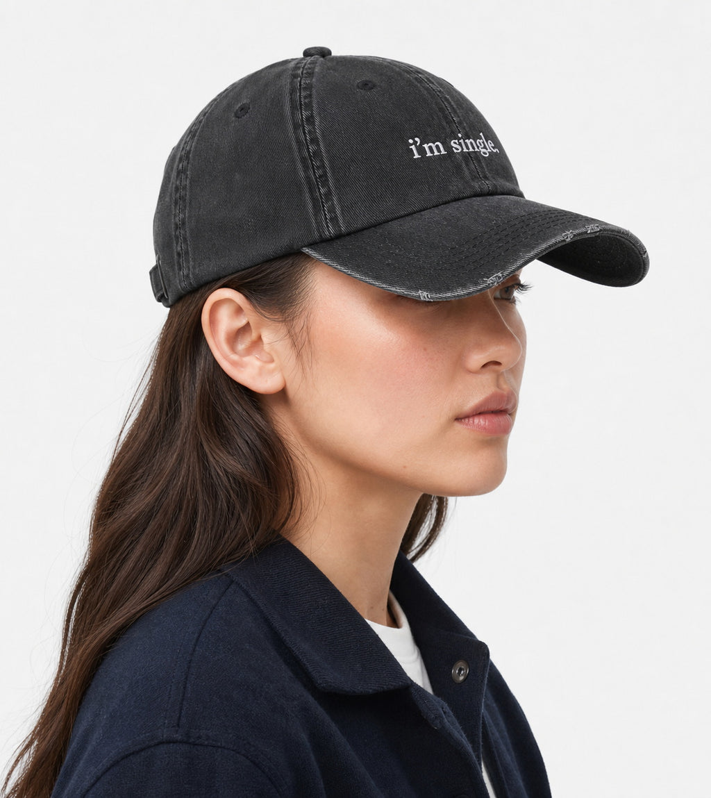 A woman in profile wearing a washed black baseball cap with the text 'i'm single' embroidered in white on the front, paired with a dark navy overshirt against a light gray background.