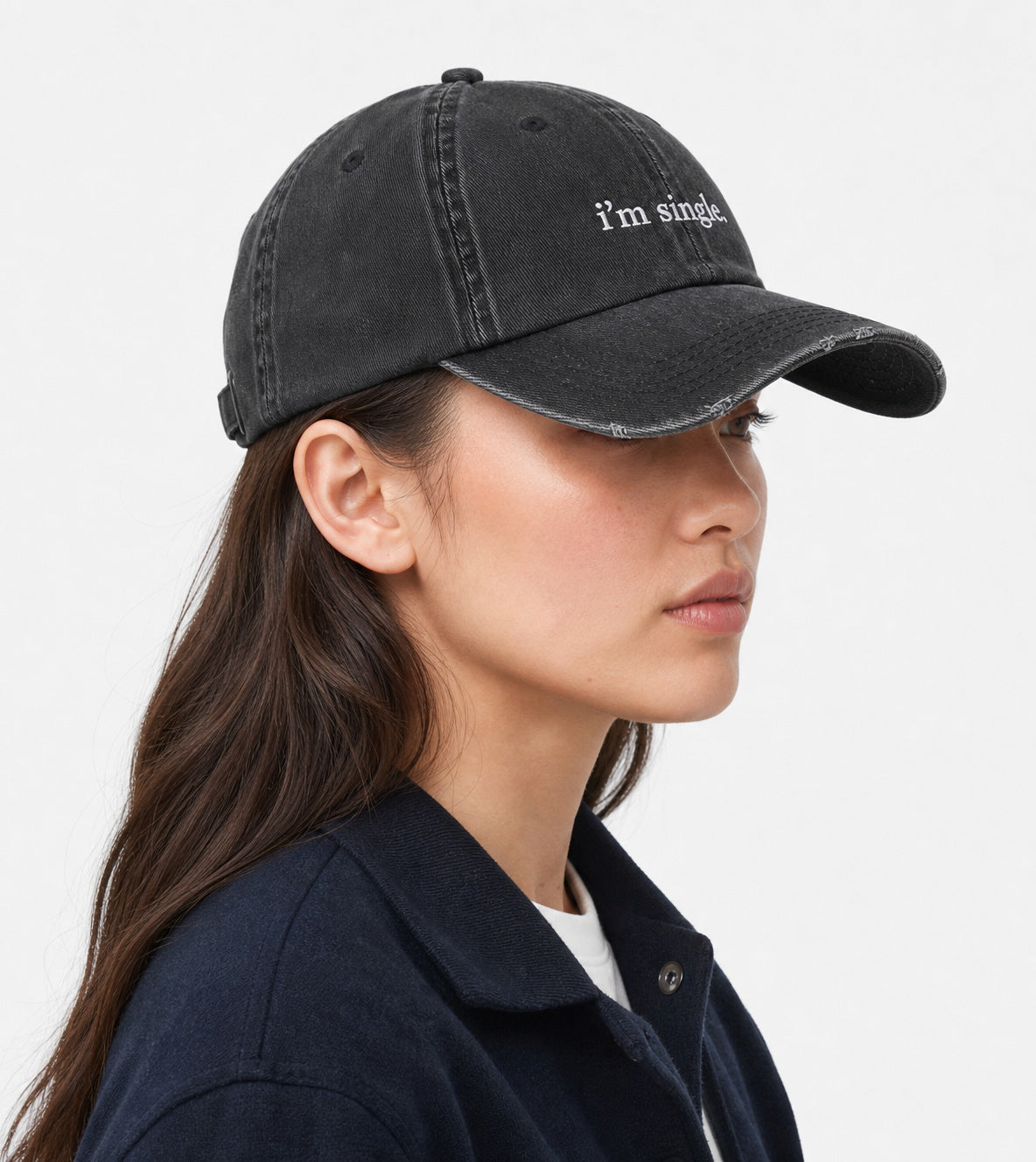 A woman in profile wearing a washed black baseball cap with the text 'i'm single' embroidered in white on the front, paired with a dark navy overshirt against a light gray background.