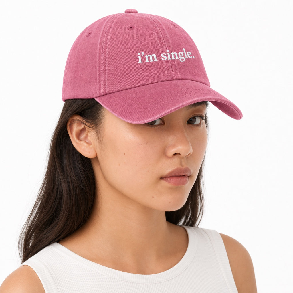 A woman wearing a dusty pink baseball cap with the text 'i'm single.' embroidered in white on the front, photographed against a clean white background.
