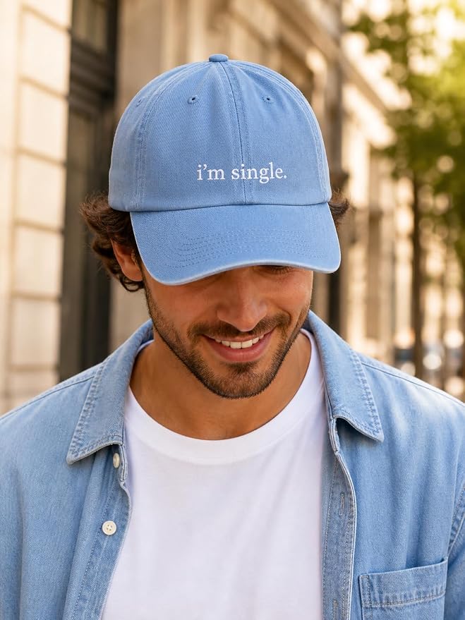 A man wearing a light blue baseball cap with the text 'i'm single.' embroidered in white on the front, styled with a white t-shirt and a light blue denim shirt in an outdoor urban setting.