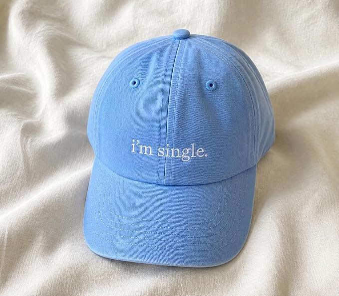 A light blue baseball cap with the phrase 'i'm single.' embroidered in white script on the front, resting on a soft, draped cream-colored fabric background.