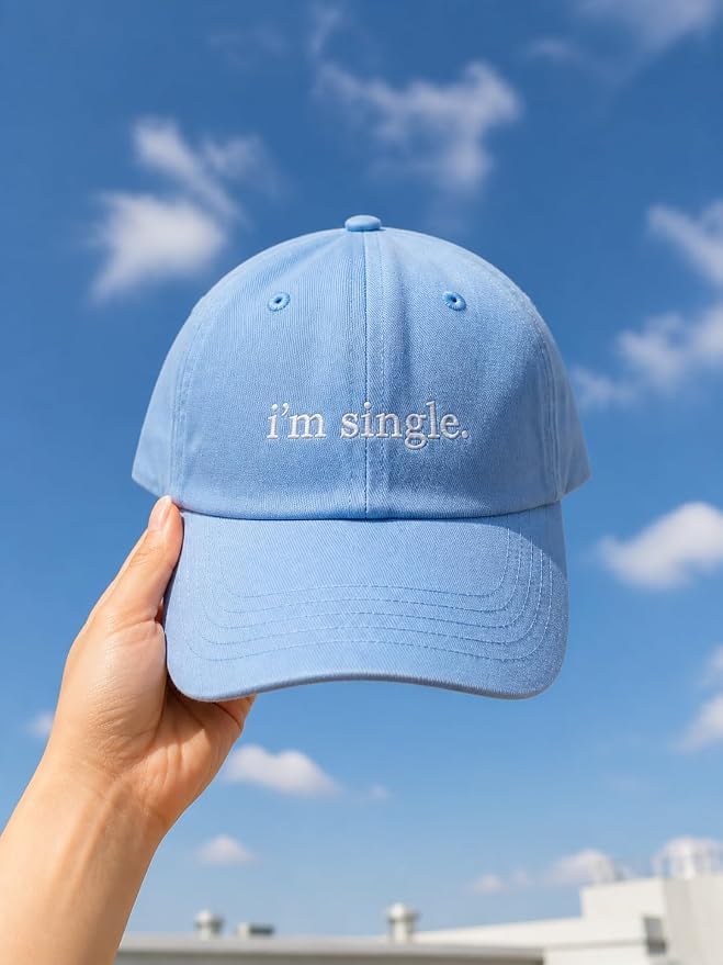 A light blue baseball cap held by a hand against a bright blue sky with white clouds. The cap features the text 'i'm single.