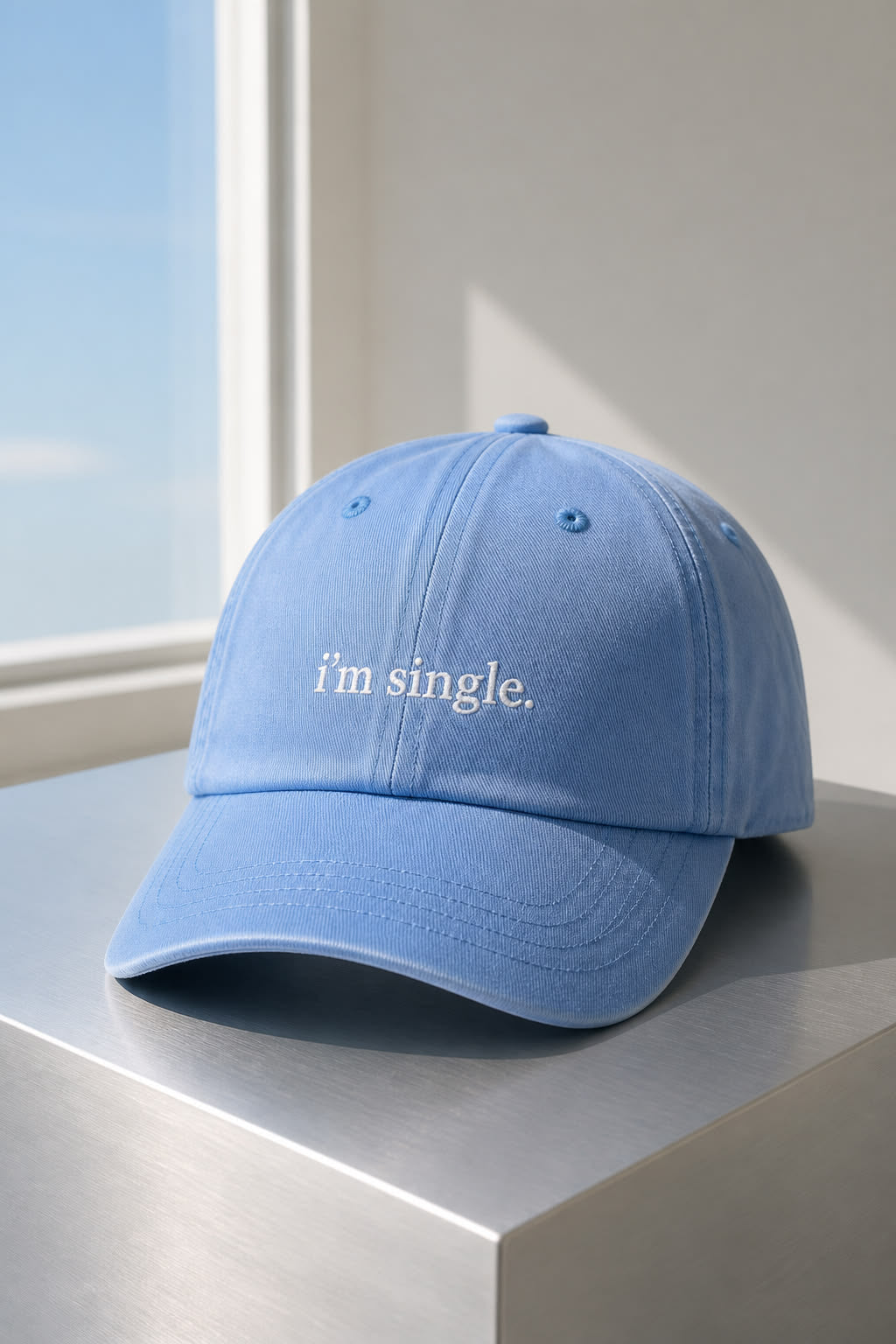 A light blue baseball cap with the phrase 'i'm single.' embroidered in white on the front, displayed on a metallic surface next to a window.