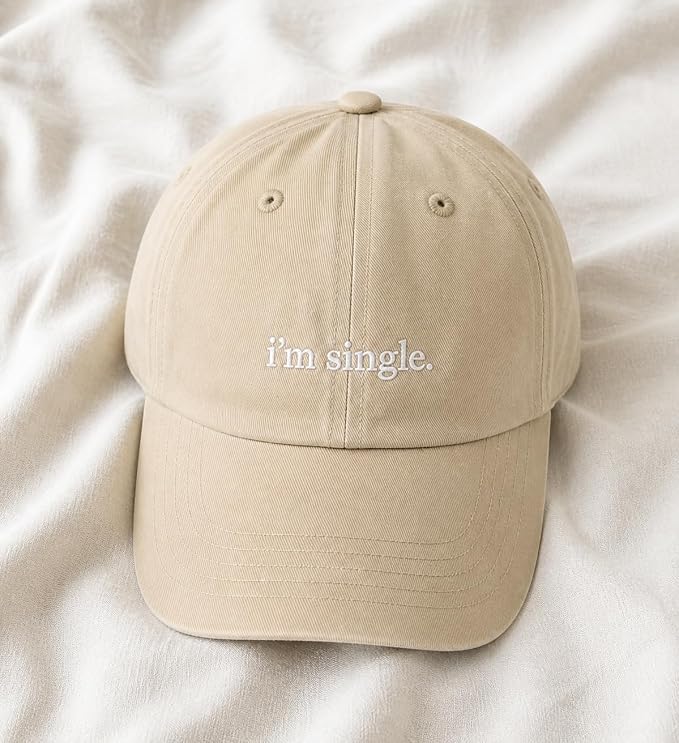 A beige baseball cap with 'i'm single.' embroidered in white on the front, resting on a soft, textured white fabric background.