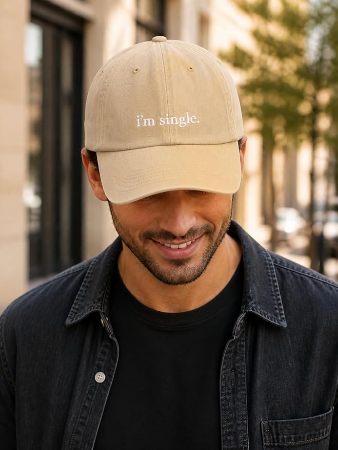 A man wearing a beige baseball cap with the text 'i'm single.' embroidered on the front, paired with a black t-shirt and a dark denim shirt jacket, set against a blurred outdoor city background.