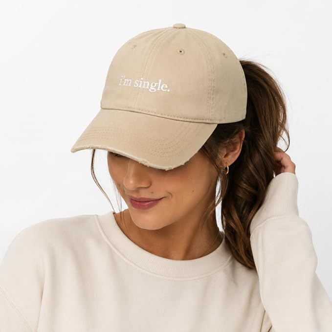 A woman wearing a beige baseball cap with the text 'I'm single.' embroidered in white on the front, paired with a cream sweatshirt against a plain white background.