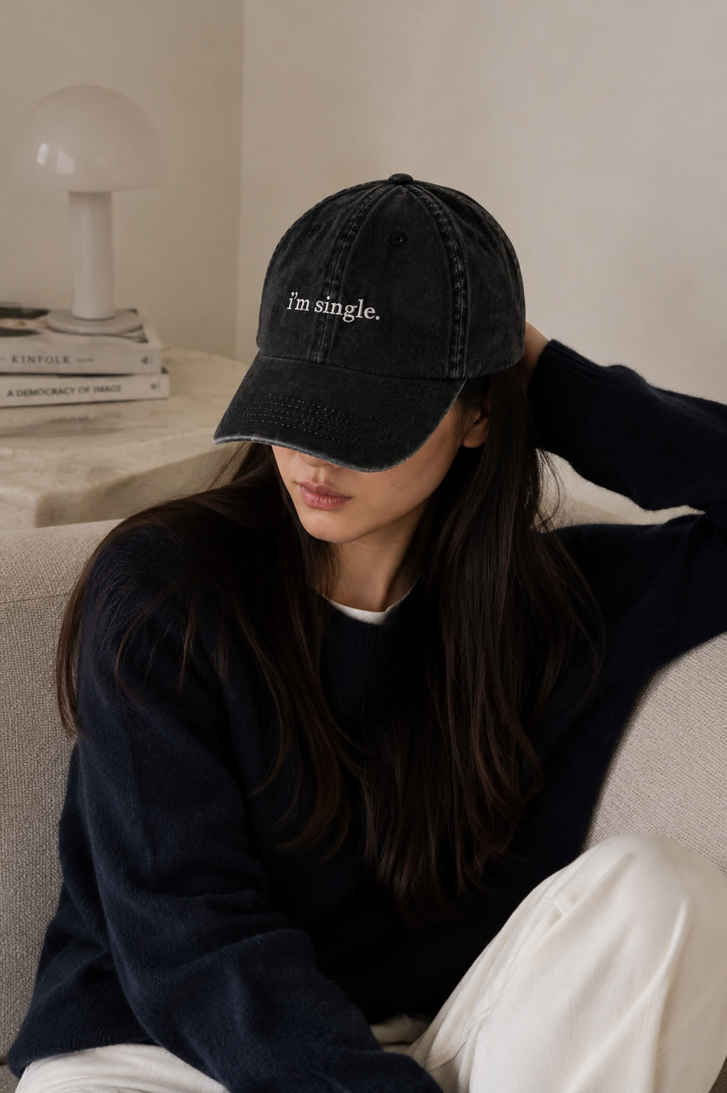 A woman wearing a black washed baseball cap with the embroidered text 'I'm single.' in white, sitting on a beige sofa in a cozy indoor setting.