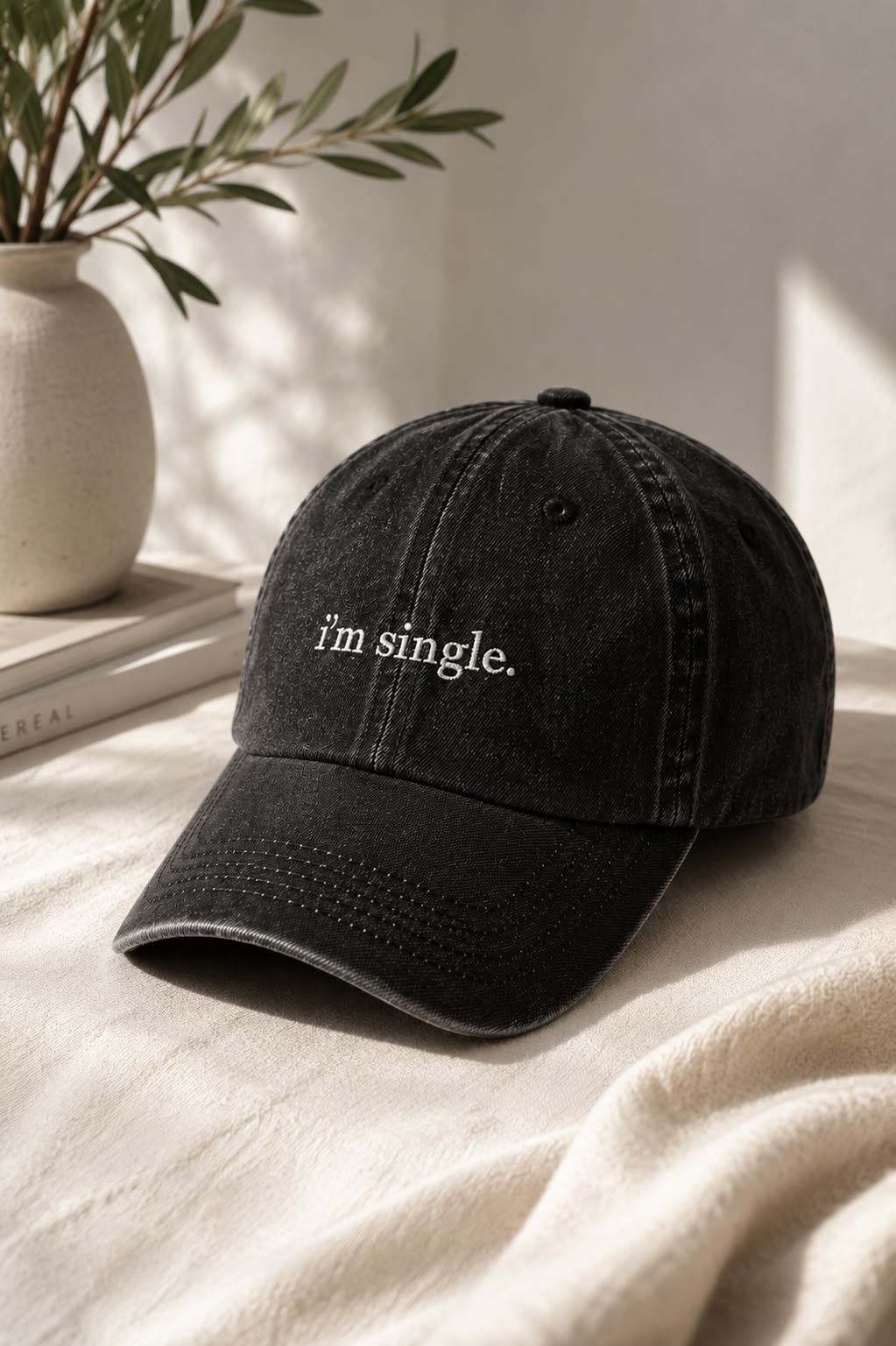 A black washed baseball cap with the phrase 'i'm single.' embroidered in white on the front, styled on a cream fabric surface next to a ceramic vase with green leaves.
