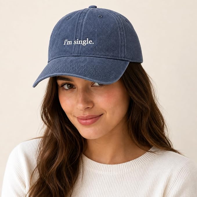 A woman wearing a blue denim baseball cap with the phrase 'i'm single.' embroidered in white on the front, set against a plain beige background.