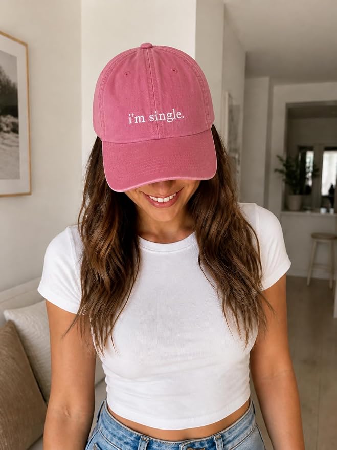 A woman wearing a dusty rose pink baseball cap with the text 'i'm single.' embroidered in white on the front, paired with a white crop top and blue jeans, standing in a brightly lit home interior.