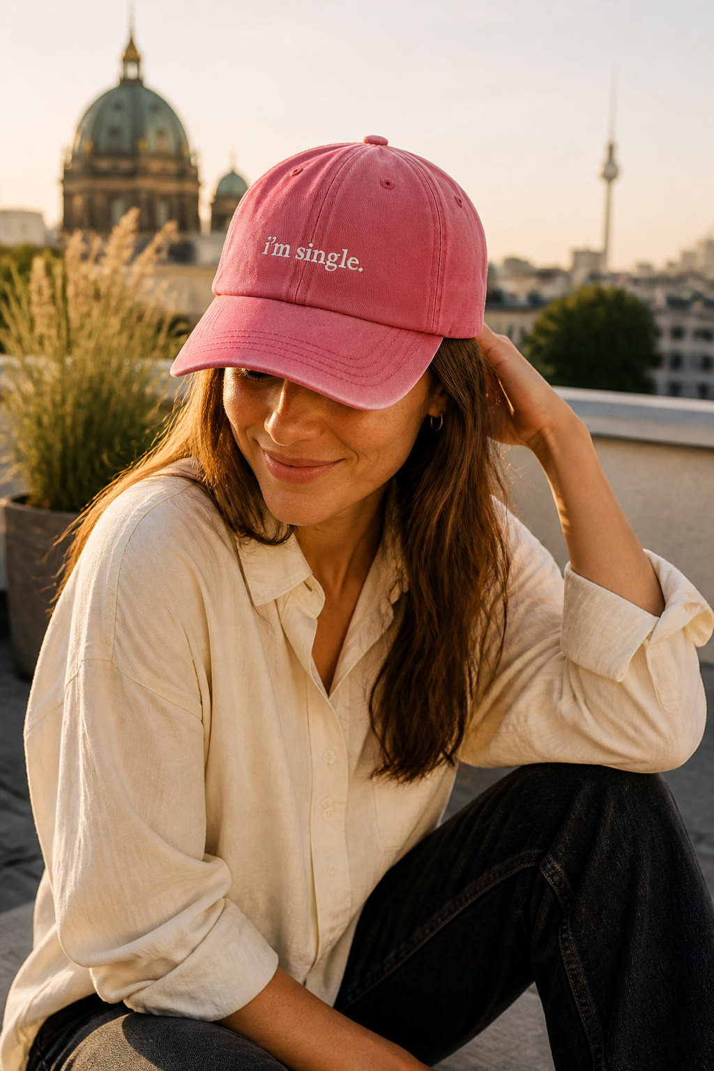 A woman sitting on a rooftop at sunset wearing a pink baseball cap with the text 'i'm single.' embroidered on the front, paired with a cream button-up shirt and dark jeans.