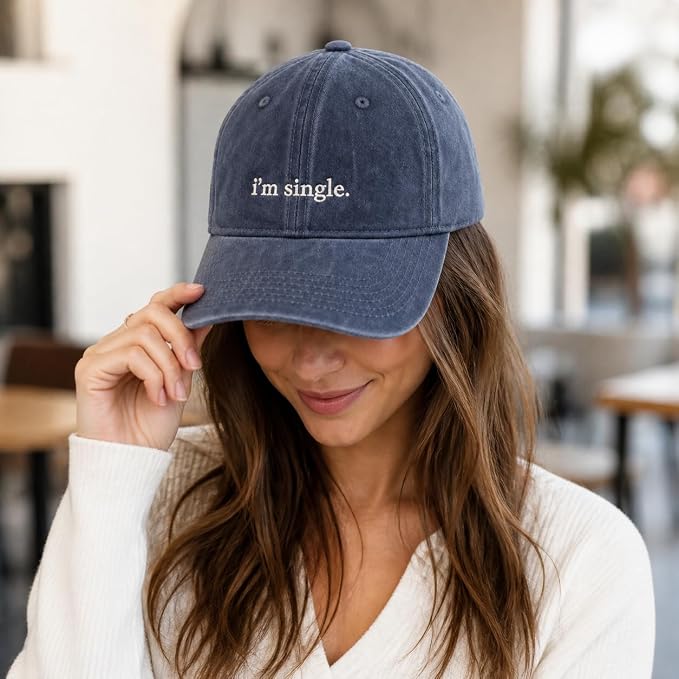 A woman wearing a washed navy blue baseball cap with the text 'i'm single.' embroidered in white on the front, holding the brim of the hat in a softly lit indoor setting.