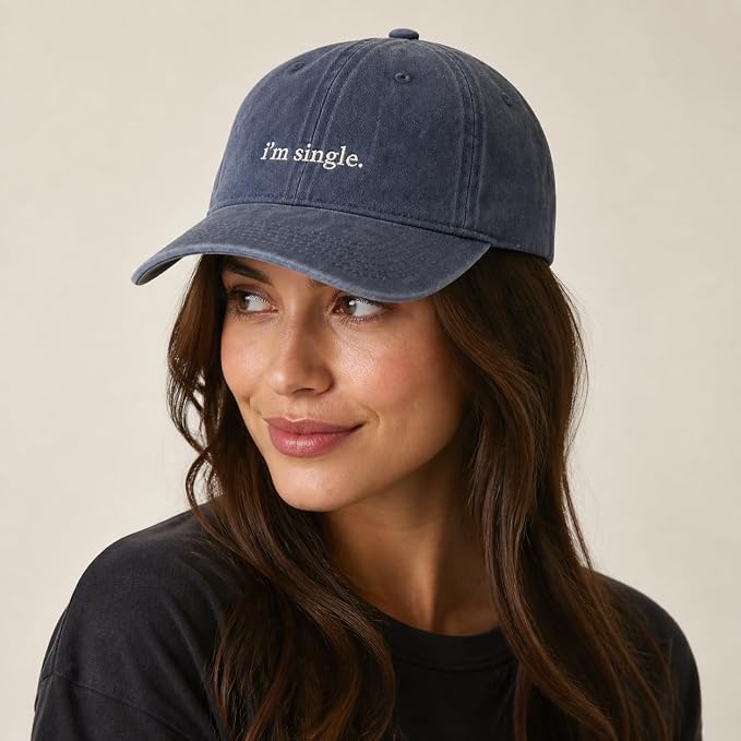 A woman wearing a washed navy blue baseball cap with the phrase 'I'm single.' embroidered in white on the front, set against a plain beige background.