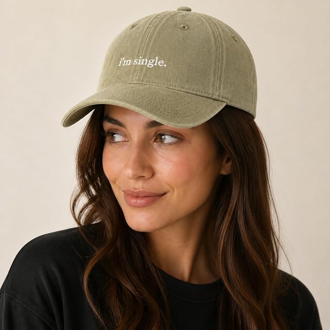 A woman wearing an olive green baseball cap with the phrase 'i'm single.' embroidered in white on the front, photographed against a plain beige background.