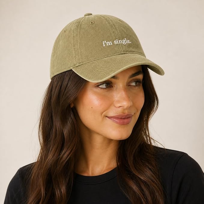 A woman wearing a washed olive green baseball cap with the text 'i'm single.' embroidered in white on the front, set against a plain beige background.