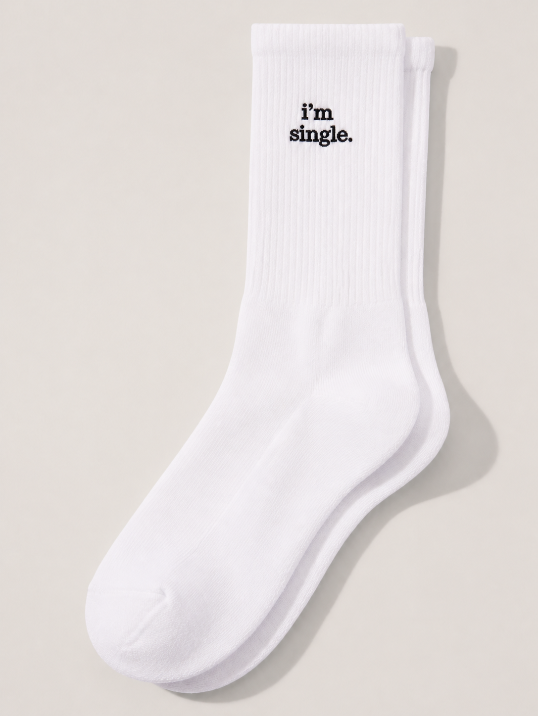 A pair of white ribbed crew socks laid flat on a cream background, with 'i'm single.' embroidered in black serif on the cuff.