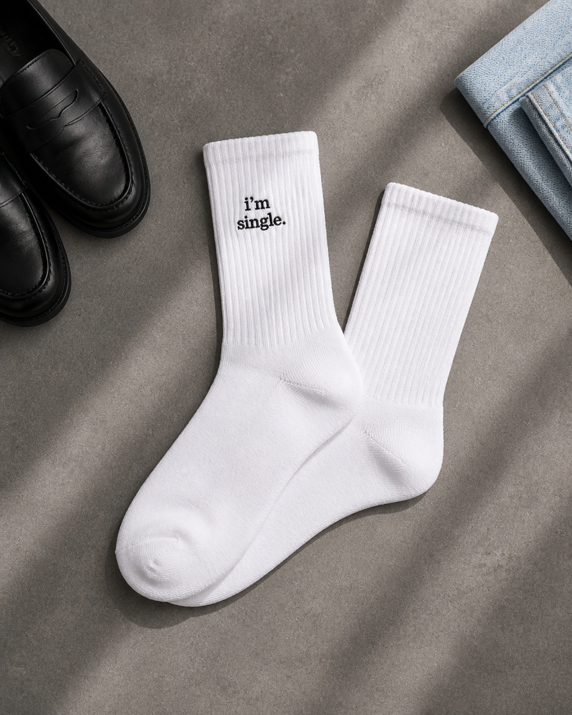 White ribbed crew socks embroidered with 'i'm single.' in black, laid flat on a concrete surface beside black leather loafers and folded light-wash denim.