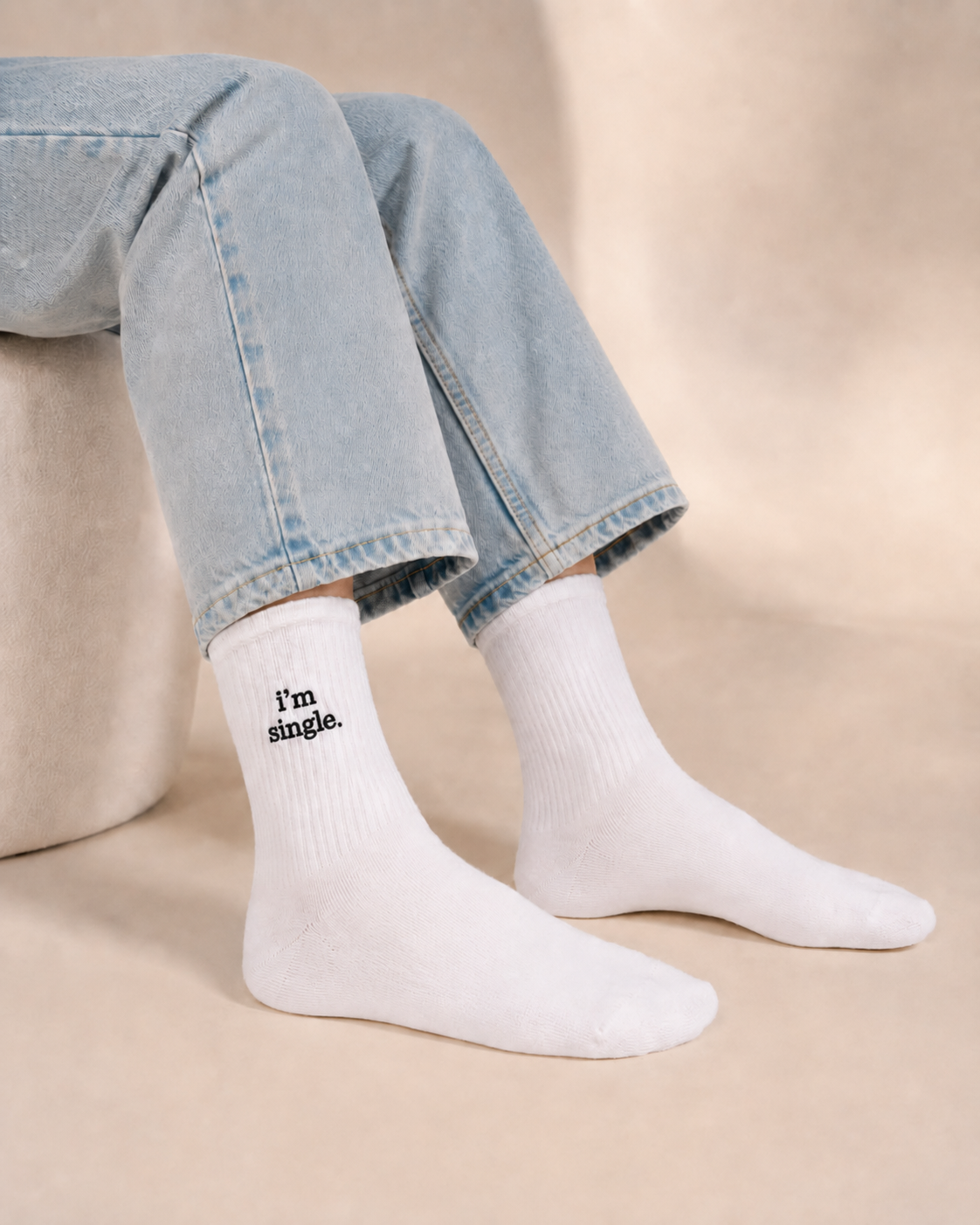 A person seated in cropped light-wash jeans wearing white ribbed crew socks embroidered with 'i'm single.' in black, against a warm beige studio background.